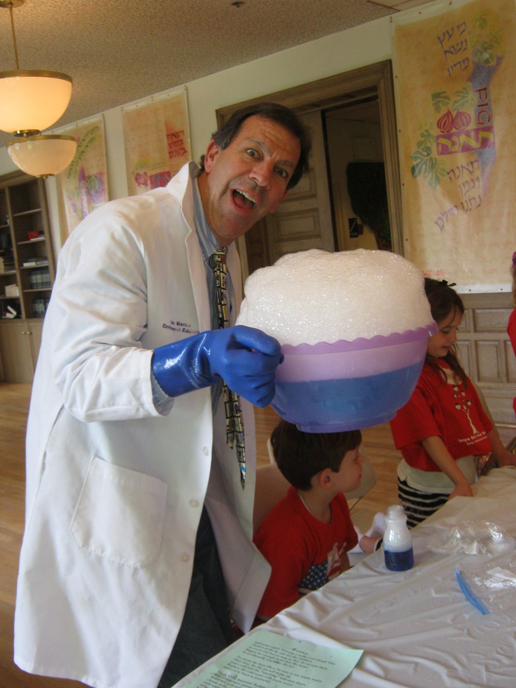 Nature Jack in a white lab coat holding a giant purple bowl overflowing with foam, smiling with preschoolers at a table