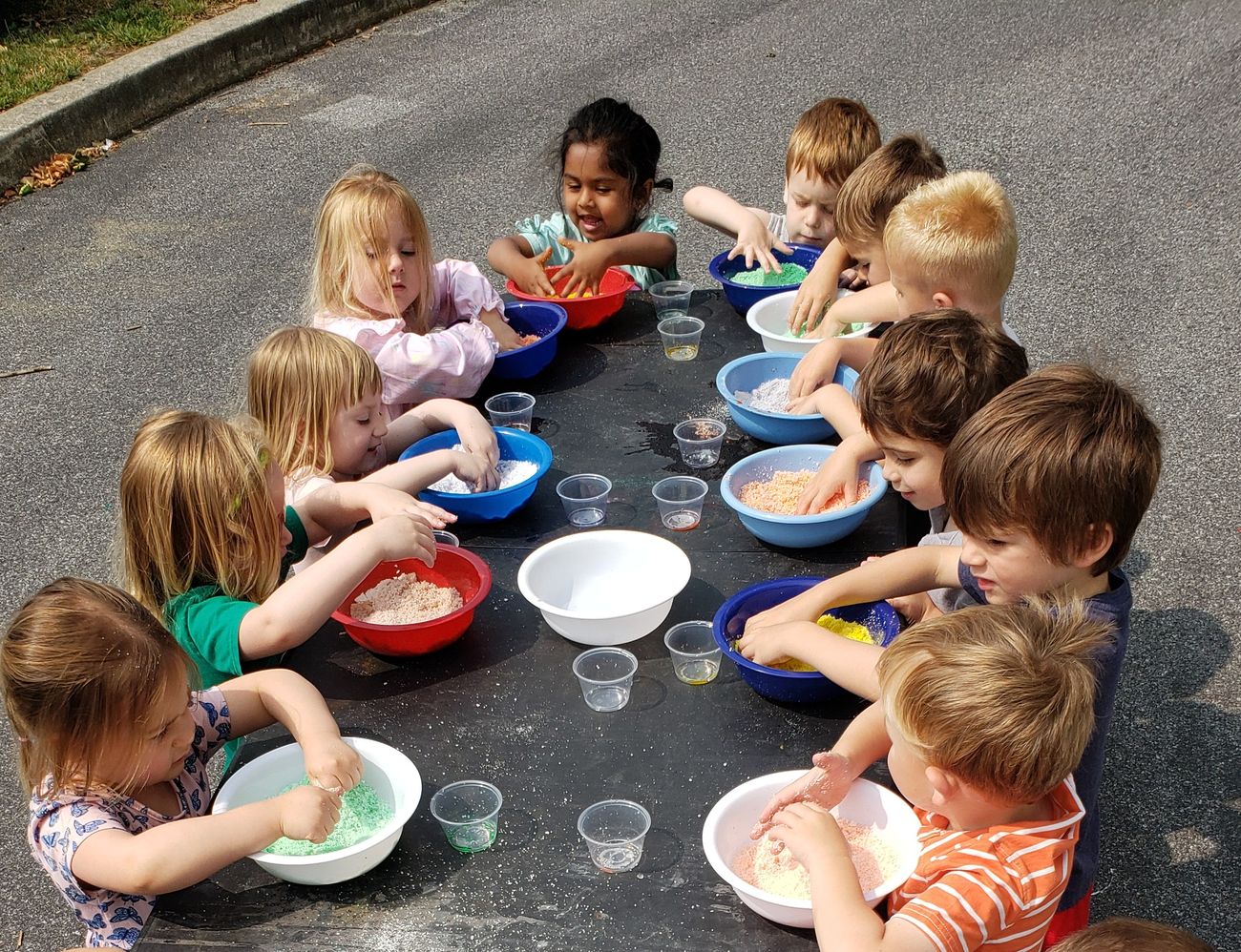 A long table of preschoolers outdoors, scooping and mixing in colorful bowls