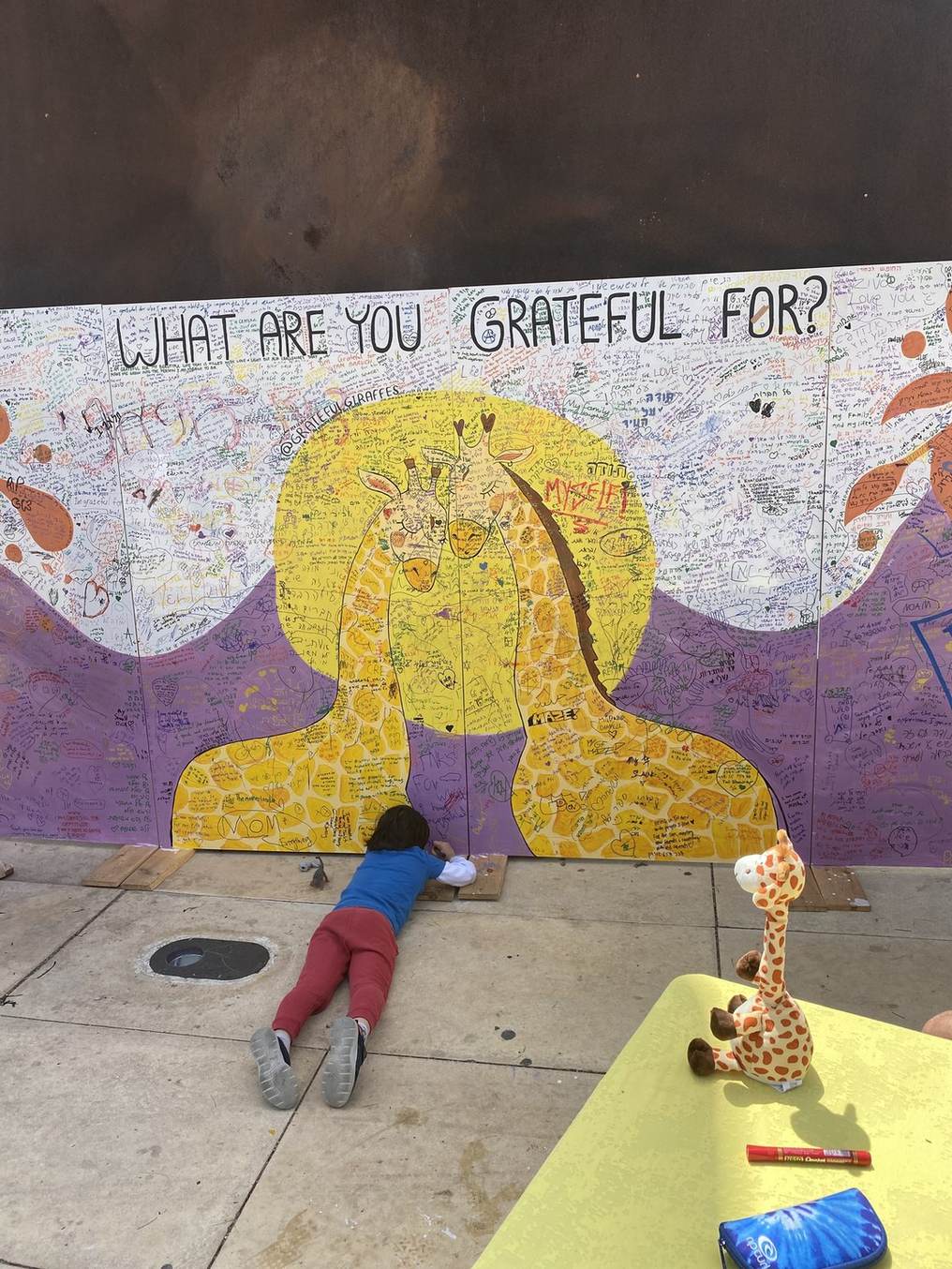 The purple and yellow Gratitude Wall in Tel Aviv, covered in handwritten notes under the words What are you grateful for, a child lying on the ground writing one more