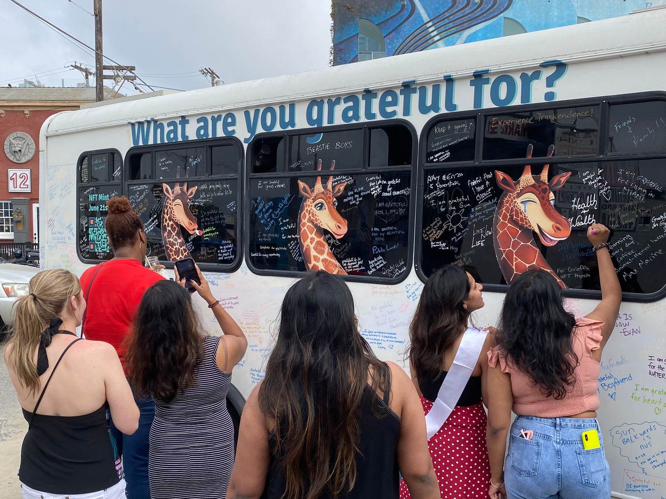 People seen from behind writing notes on the side of the white Grateful Bus, under the words What are you grateful for and painted giraffes in its windows