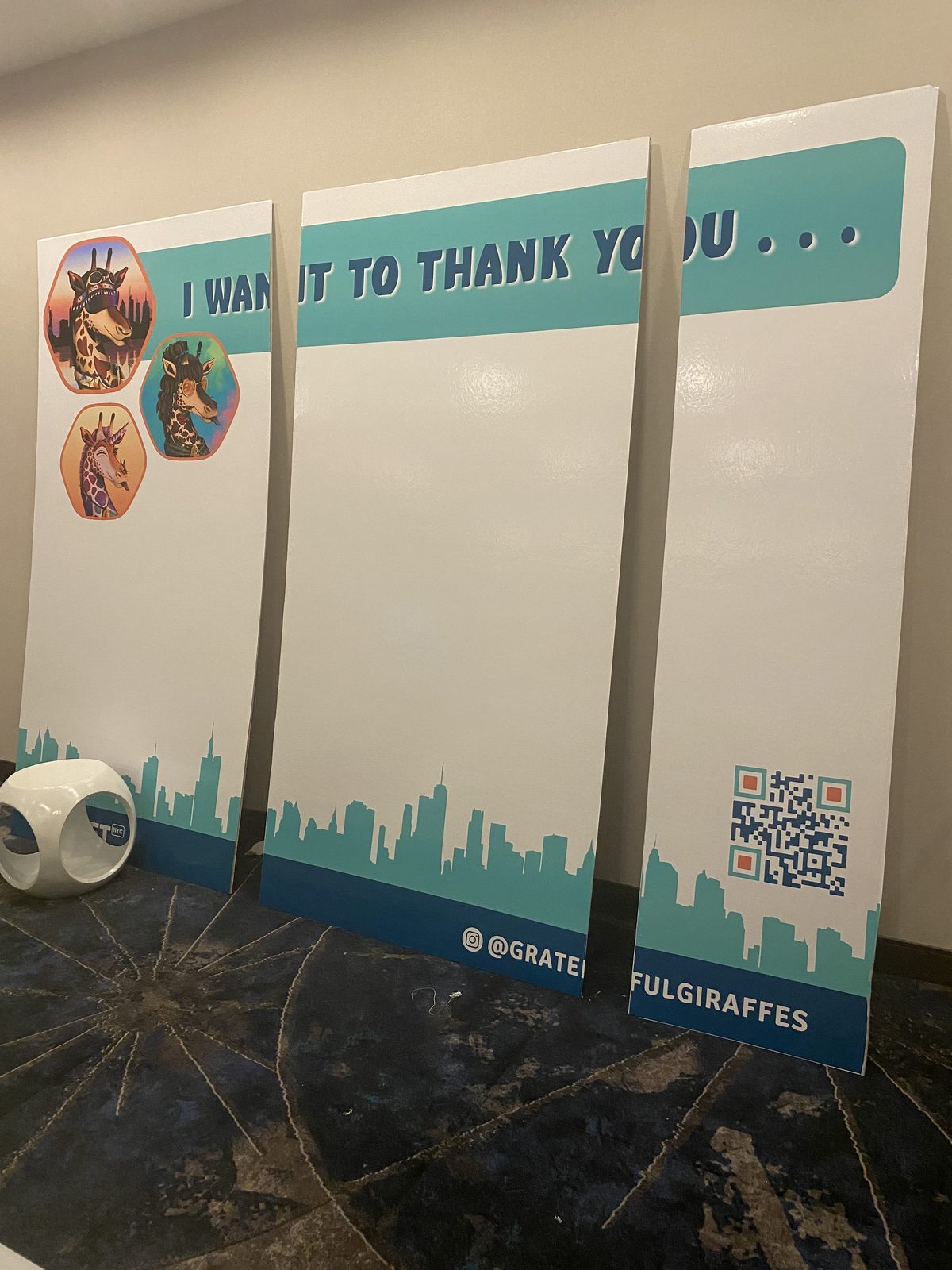 Three blank white panels headed I want to thank you, with giraffe portraits and a QR code, leaning against a wall on a hotel carpet