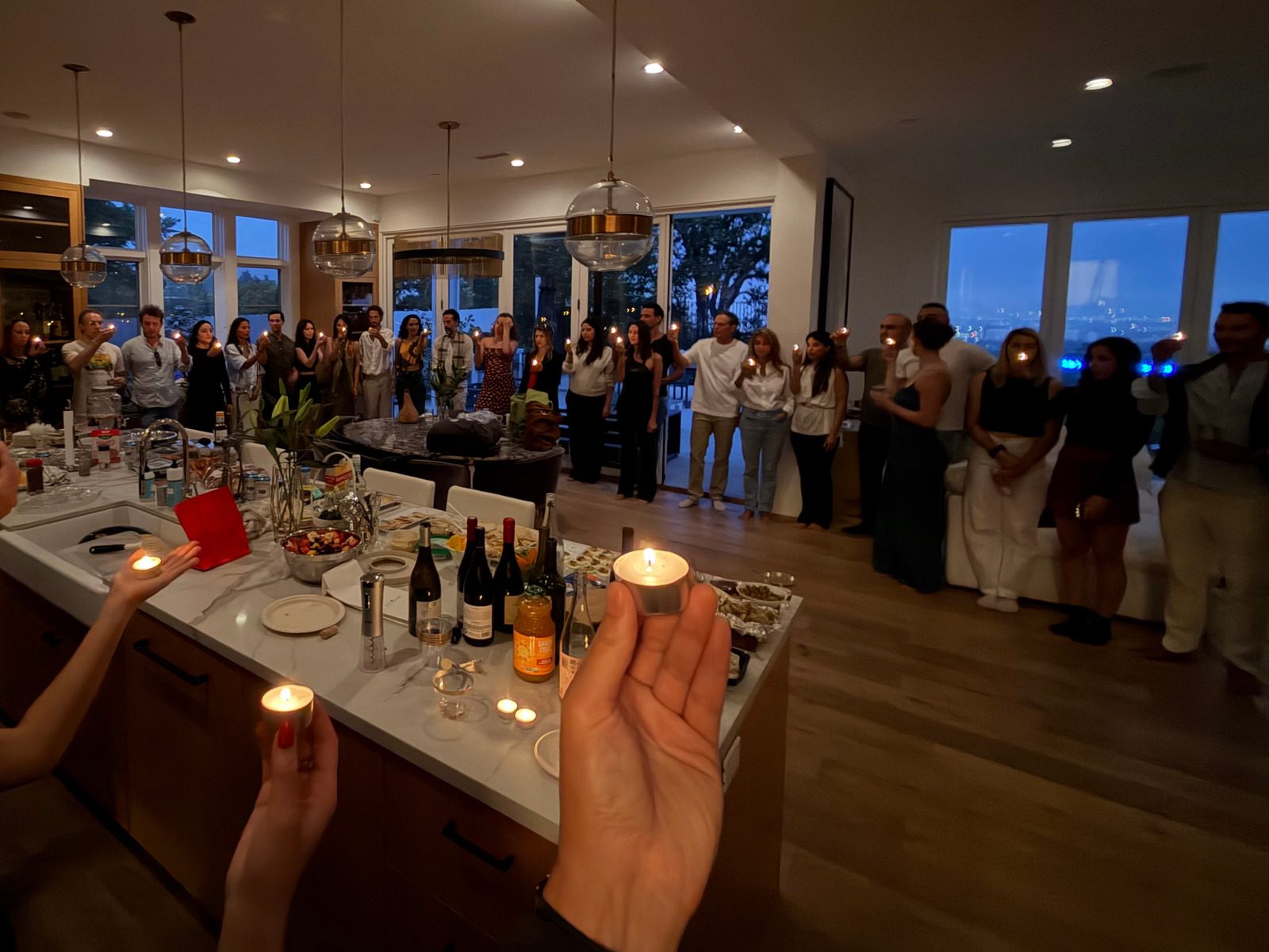 About thirty-five Giraffes standing in a wide circle around a living room at dusk, each holding a lit tea candle, two hands with candles in the foreground